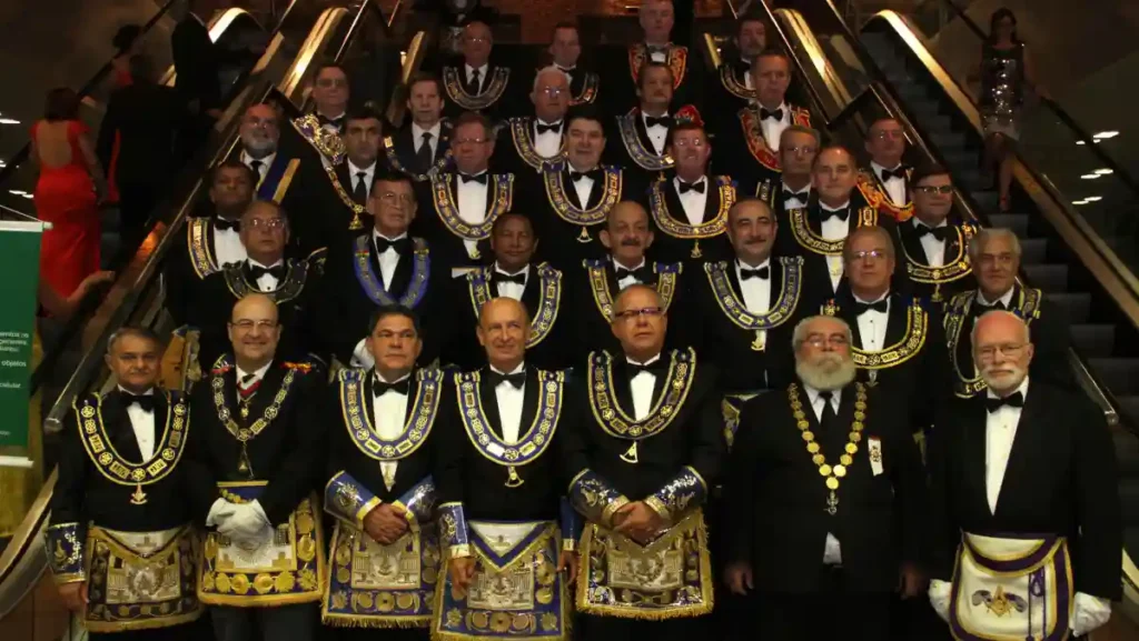 Homens usando trajes formais e paramentos da maçonaria durante cerimônia maçônica em ambiente fechado