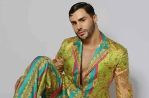 Modelo Agustín Fernandes posando sentado com expressão séria, usando terno estampado multicolorido em ensaio fotográfico de moda.