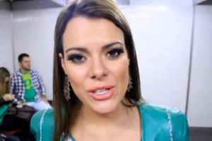Cantora gospel falando em bastidores de evento, com maquiagem destacada e figurino azul, enquanto pessoas aparecem desfocadas ao fundo.