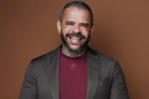 Pastor sorrindo em retrato profissional, usando blazer escuro e camiseta vinho diante de fundo marrom.