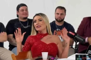 Mulher sentada em estúdio levantando as mãos durante entrevista, usando vestido vermelho, com microfone e outras pessoas ao fundo.
