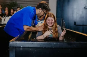 Atriz emocionada durante batismo em tanque de imersão enquanto líder cristão a acompanha colocando a mão em seu ombro.