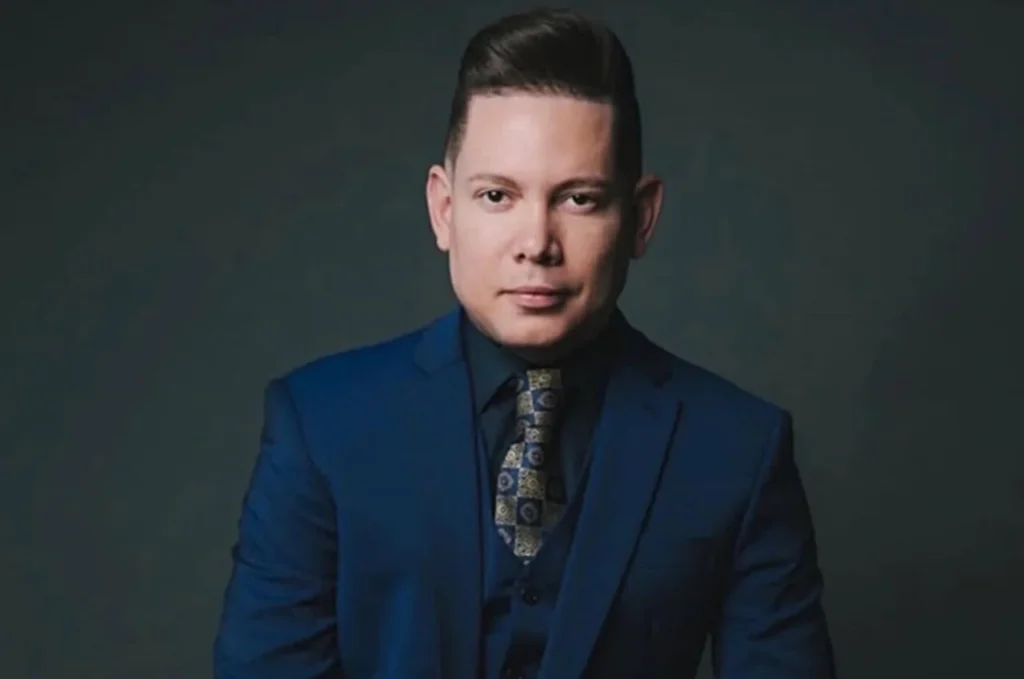 Homem de terno azul posando para retrato com expressão séria em fundo escuro.