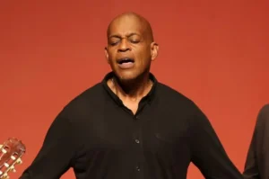 Cantor gospel de olhos fechados, cantando com expressão emocional diante de fundo vermelho.