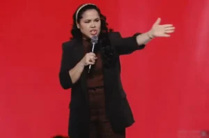 Cassiane pregando com expressão firme e microfone na mão durante culto em auditório com fundo vermelho.