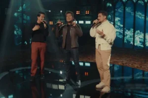 Cantor Daniel se apresenta ao vivo ao lado de dois músicos, segurando microfone e gesticulando no palco iluminado durante gravação musical.