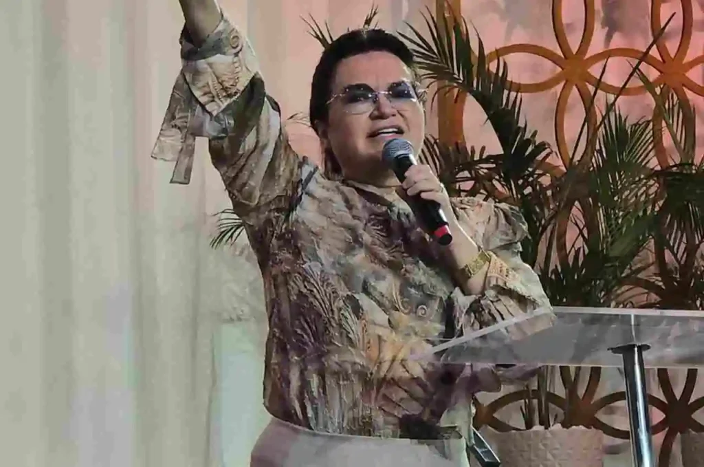 Pastora pregando no púlpito com uma das mãos erguidas e microfone na outra, diante de decoração com plantas e iluminação suave.