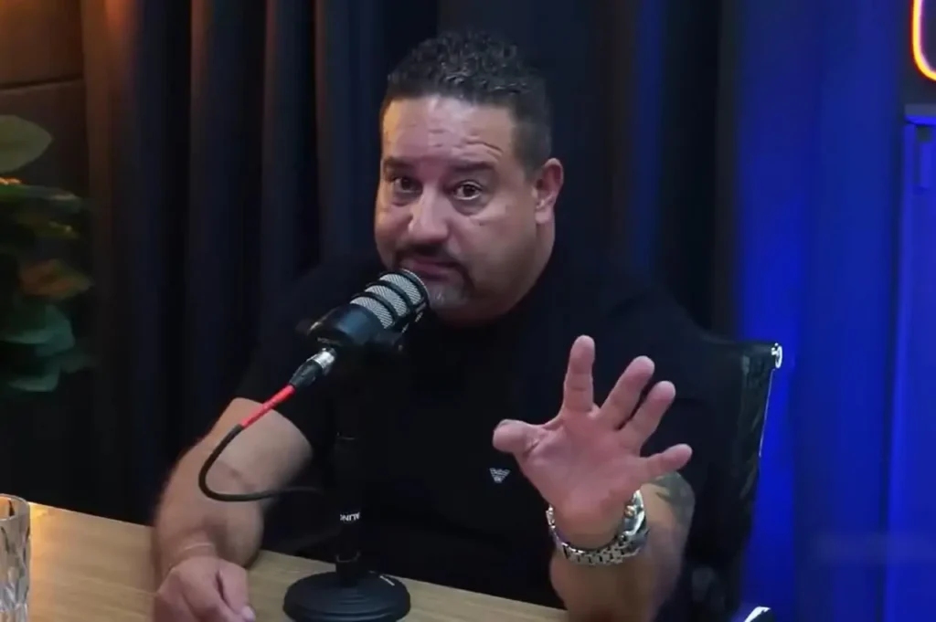 Pastor falando em mesa de podcast, gesticulando com a mão enquanto utiliza microfone profissional em estúdio iluminado.