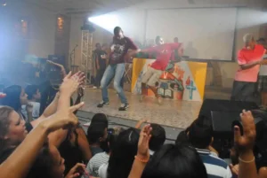Jovens dançando em apresentação de música urbana em evento com público cristão, com plateia animada e palco iluminado.