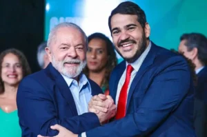 Lula e Messias sorrindo e apertando as mãos durante evento oficial, posando juntos em frente a painel institucional.