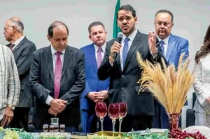 Autoridades reunidas em cerimônia de cunho religioso, com um homem de terno falando ao microfone enquanto outros permanecem em postura de oração diante de mesa com cálices e elementos simbólicos.