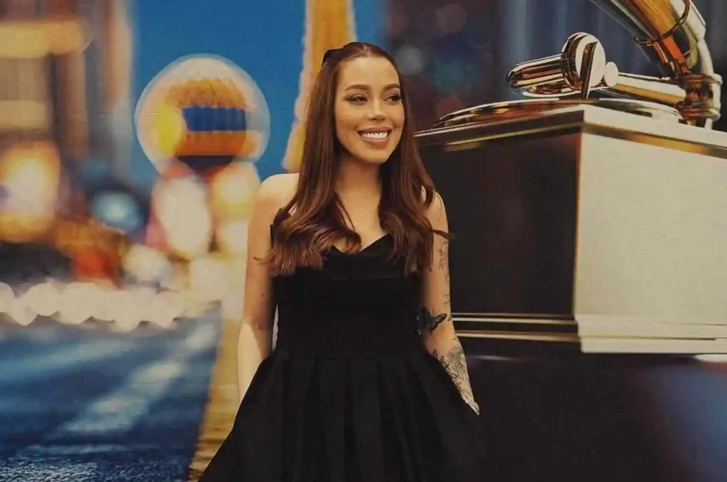 Cantora sorrindo enquanto posa com vestido preto diante de painel iluminado com elemento cenográfico dourado semelhante a troféu musical.