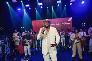 Homem negro usando terno branco canta com microfone em um palco iluminado, acompanhado por banda e coral ao fundo.
