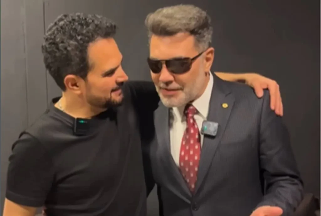 Famosos conversando de forma descontraída, abraçados, sendo um deles trajado com terno e óculos escuros, enquanto o outro usa camiseta preta com microfone de lapela.