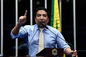 Parlamentar discursando em plenário, gesticulando com o dedo levantado enquanto fala ao microfone, com bandeira nacional ao fundo.