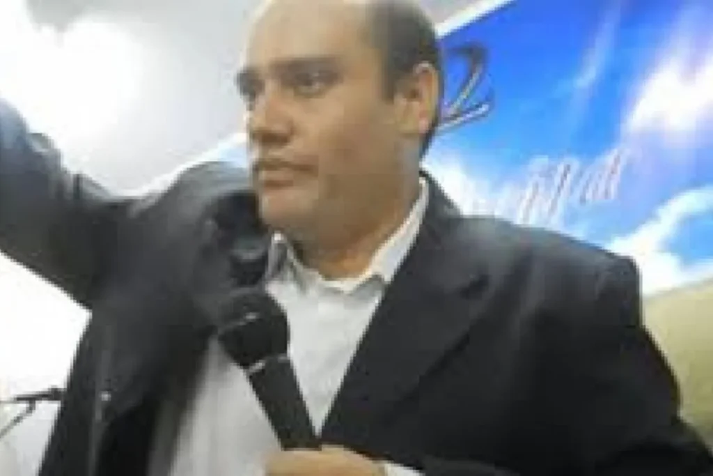 Pastor segurando microfone enquanto gesticula durante pregação em ambiente de culto com painel azul ao fundo.