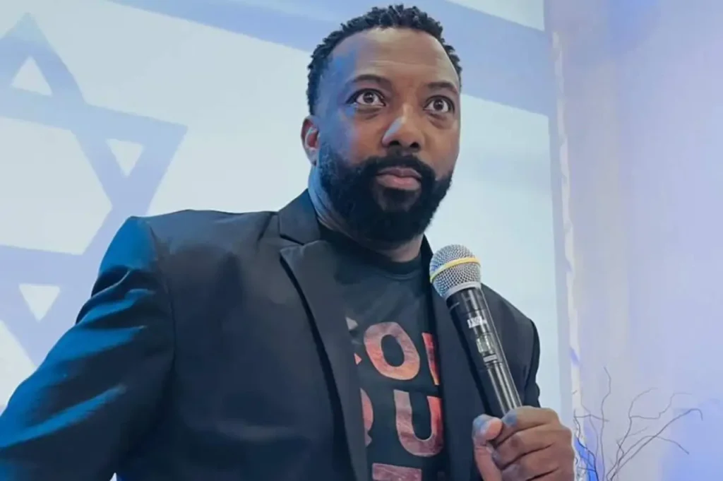 Pastor segurando microfone enquanto prega em ambiente interno iluminado, com símbolo religioso estilizado ao fundo.