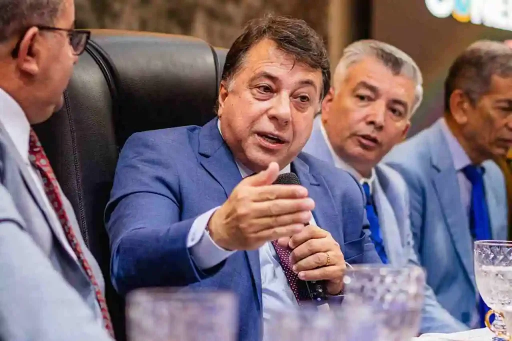 Pastor sentado à mesa durante reunião ministerial, falando ao microfone com gesto enfático enquanto outros líderes observam ao fundo.