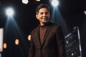 Pastor Jairo Manhães sorrindo no palco durante evento, sob iluminação profissional.