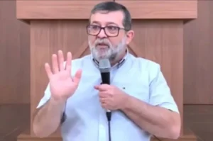 Pastor segura microfone e faz gesto com a mão durante pregação em igreja.