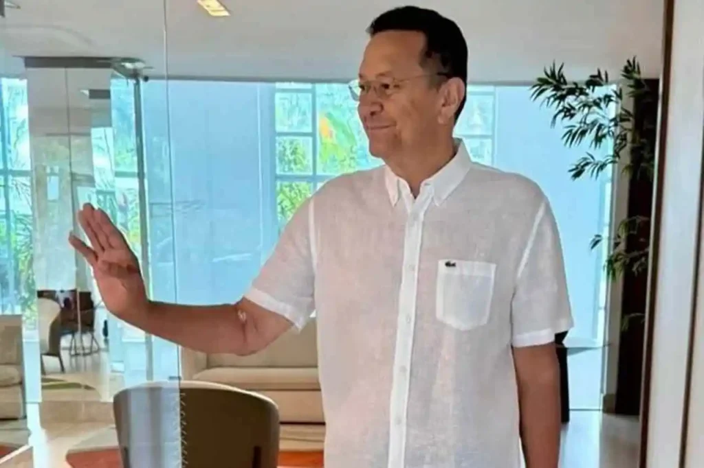 Pastor em ambiente interno acenando com a mão, vestido com camisa social clara e expressão serena.