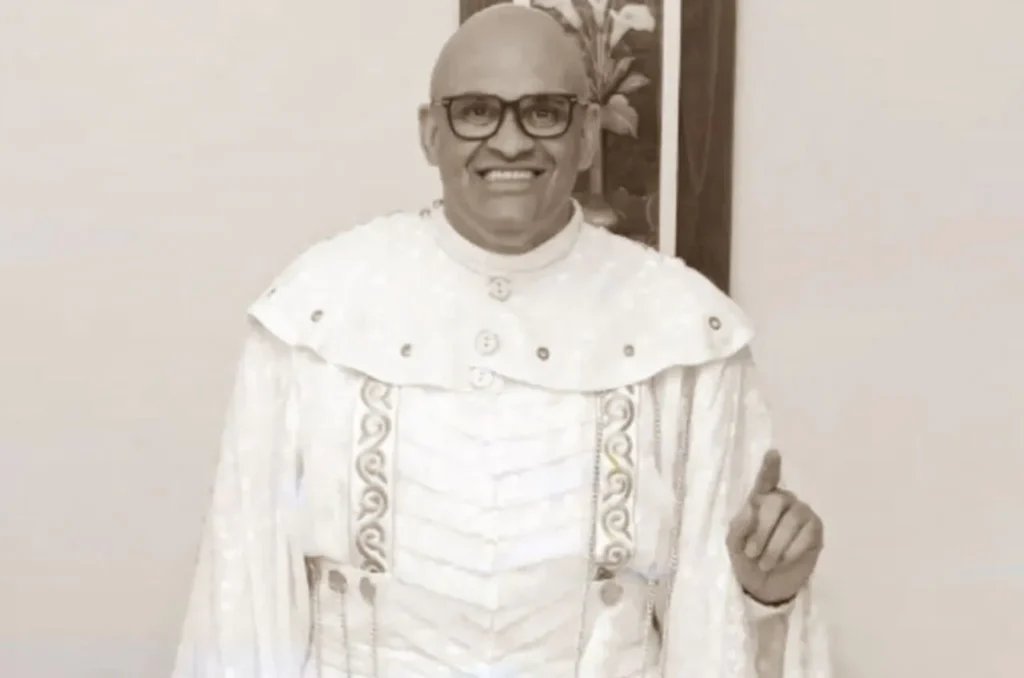Pastor vestindo túnica litúrgica branca, sorrindo e fazendo gesto com o dedo levantado em ambiente interno.