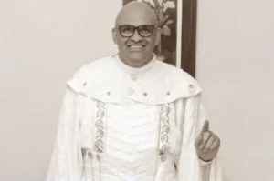 Pastor vestindo túnica litúrgica branca, sorrindo e fazendo gesto com o dedo levantado em ambiente interno.