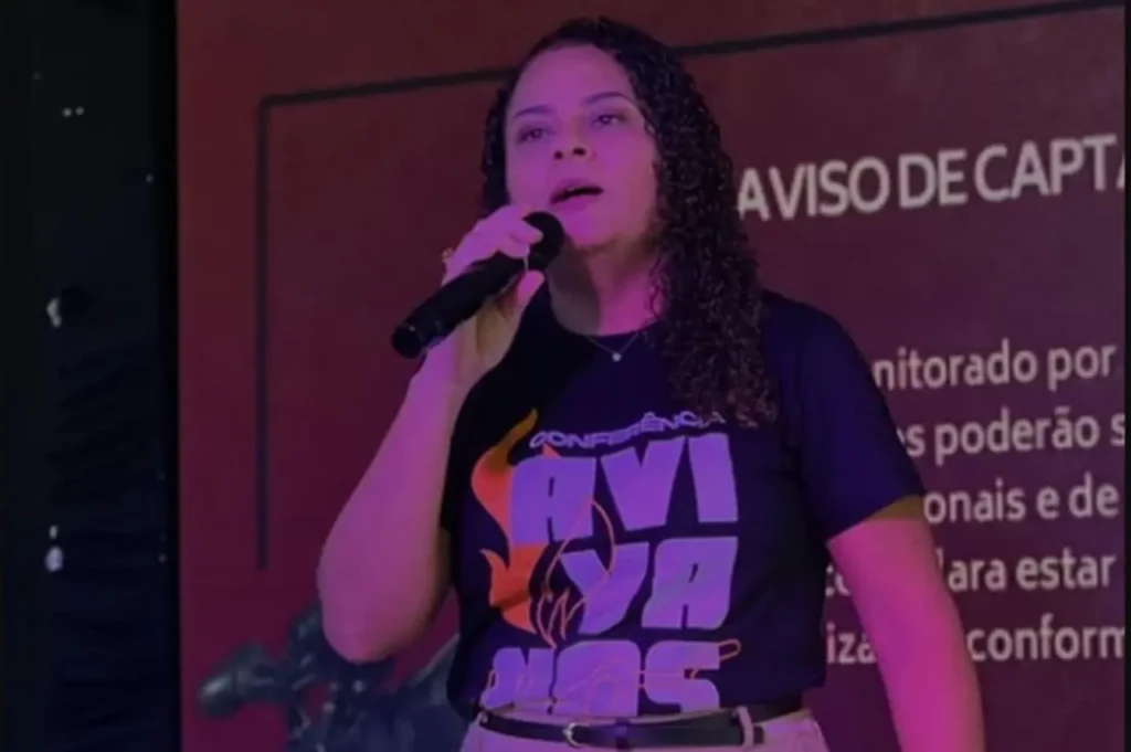 Líder religiosa segurando microfone e ministrando durante conferência evangélica, em palco iluminado com painel ao fundo.