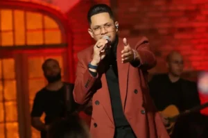 Cantor gospel se apresentando ao vivo no palco, segurando microfone e apontando para o público, com iluminação cênica em tons quentes ao fundo.