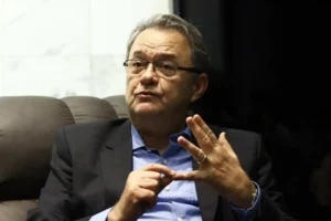 Pastor sentado em poltrona gesticulando enquanto fala, usando terno escuro e camisa azul durante entrevista em ambiente interno.