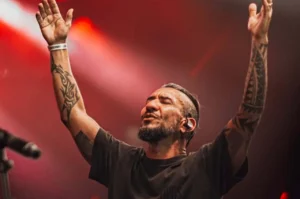 Homem tatuado, de barba e cabelo preso, levanta os braços e canta de olhos fechados em um palco iluminado por luz vermelha.