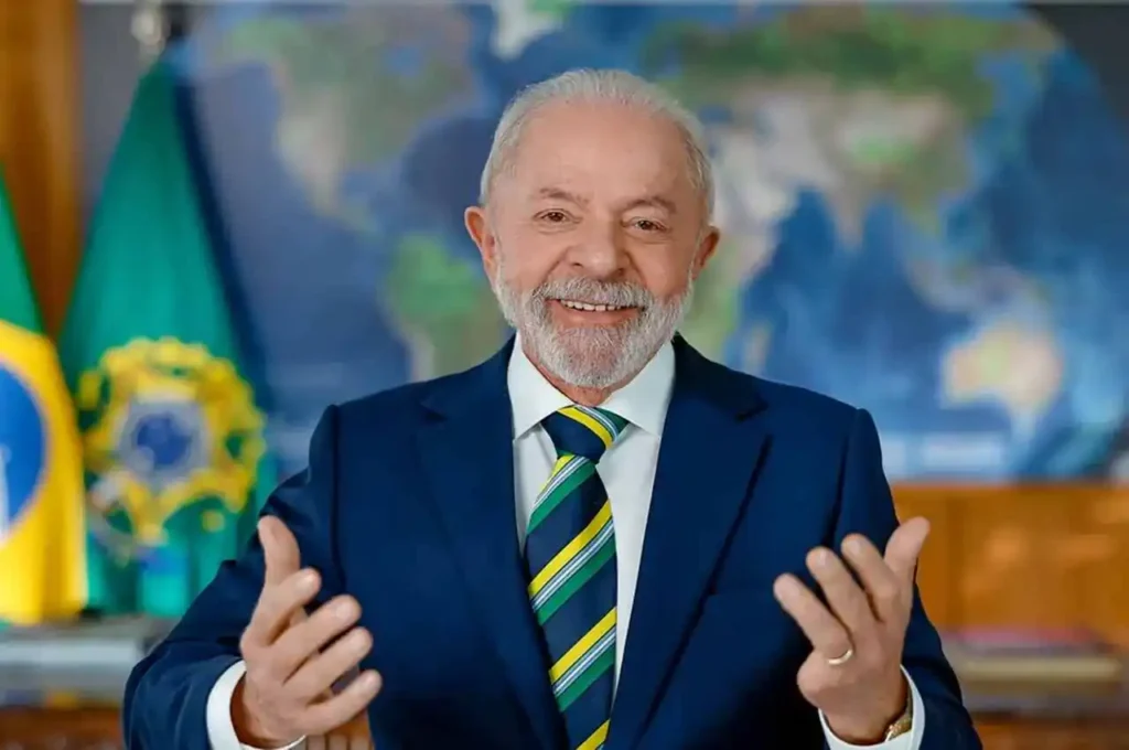 Lula usando terno azul e gravata com listras verdes e amarelas, fala sorrindo com as mãos erguidas diante de bandeiras e um mapa ao fundo.