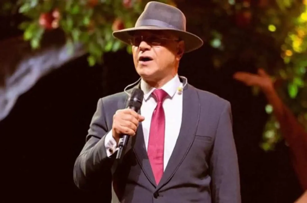 Cantor Sérgio Lopes no palco, usando terno escuro, gravata vermelha e chapéu, segurando um microfone durante apresentação.