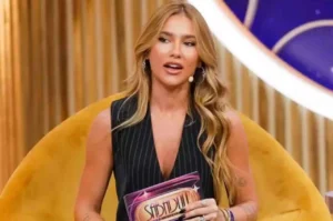 Apresentadora loira sentada em poltrona amarela segurando ficha de programa de TV.