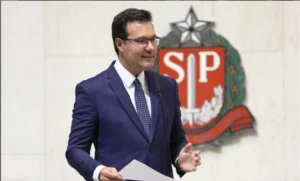 Deputado estadual Alex de Madureira discursando na tribuna da Alesp, com brasão de São Paulo ao fundo