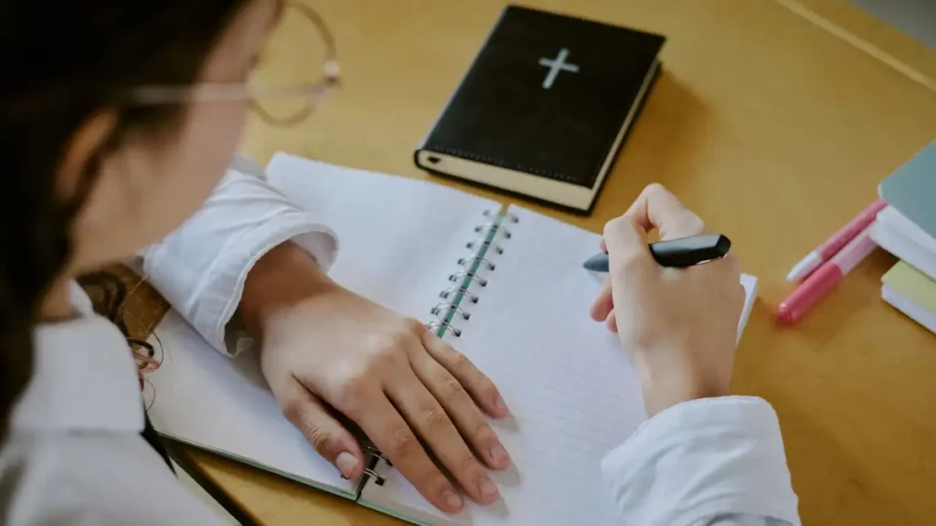 pessoa fazendo anotacoes em caderno enquanto estuda a biblia sobre mesa de madeira