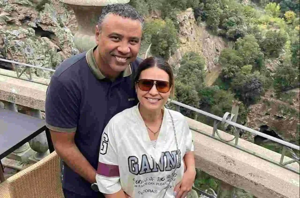 Aline Barros posa sorridente ao lado do marido, Gilmar, em passeio turístico ao ar livre, com paisagem rochosa e área verde ao fundo.