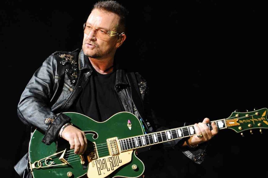 Bono Vox, vocalista do U2, tocando guitarra elétrica verde durante apresentação ao vivo no palco, com iluminação escura ao fundo.