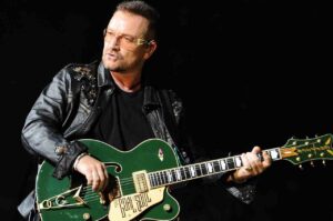 Bono Vox, vocalista do U2, tocando guitarra elétrica verde durante apresentação ao vivo no palco, com iluminação escura ao fundo.