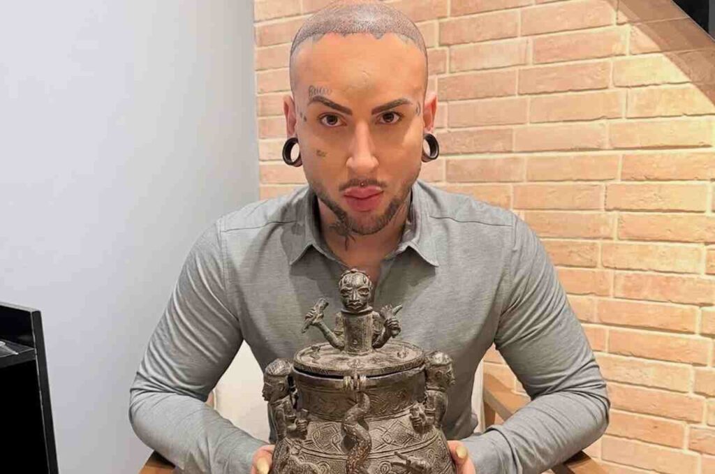 Bruxo Malagueta exibindo artefato ritualístico com símbolos esotéricos sobre mesa, em ambiente interno com parede de tijolos aparentes.