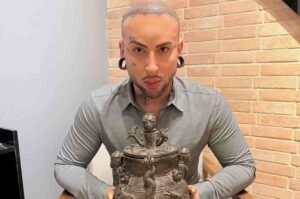 Bruxo Malagueta exibindo artefato ritualístico com símbolos esotéricos sobre mesa, em ambiente interno com parede de tijolos aparentes.