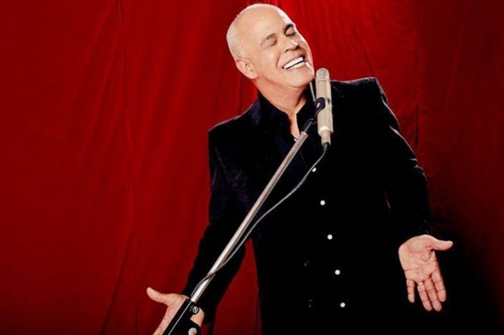 Cantor Mattos Nascimento interpretando louvor com sorriso e braços abertos diante de microfone, em ensaio ou apresentação com fundo vermelho.