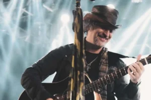 Cantor PG tocando violão e cantando com expressão de alegria durante apresentação ao vivo em show gospel.