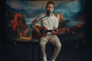 O cantor Rodrigo Siqueira canta com os olhos fechados enquanto toca violão, em apresentação acústica com microfone e cenário artístico ao fundo.