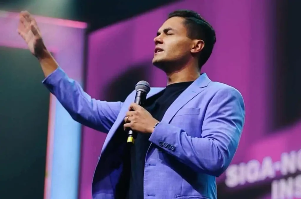 Pastor Davi Miranda Neto ministrando com mão erguida e microfone no palco durante culto em igreja evangélica.