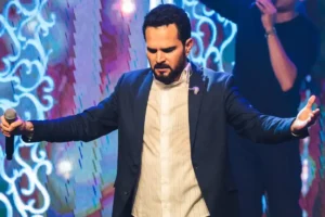 Cantor Luciano em apresentação ao vivo, de terno escuro, com braços abertos e microfone no palco iluminado.