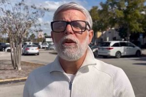 O pastor Cláudio Duarte aparece ao ar livre, usando óculos e barba grisalha, em área urbana durante gravação ou pronunciamento.