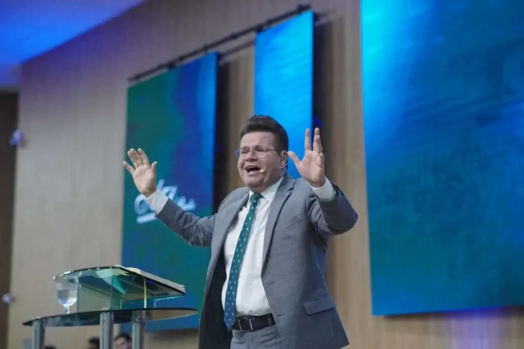 Jacques Balbino ministra mensagem com as mãos erguidas em púlpito de igreja, durante culto, com painéis iluminados ao fundo.
