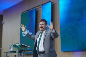 Jacques Balbino ministra mensagem com as mãos erguidas em púlpito de igreja, durante culto, com painéis iluminados ao fundo.