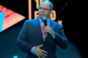 Pastor Josué Gonçalves pregando com microfone na mão durante evento cristão, usando terno azul e expressão concentrada no palco.